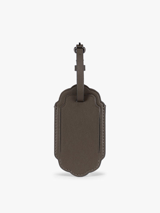 A dark brown CALPAK Vault luggage tag with a scalloped border and a matching dark brown strap, shown against a plain white background; ATV2601-MUSHROOM