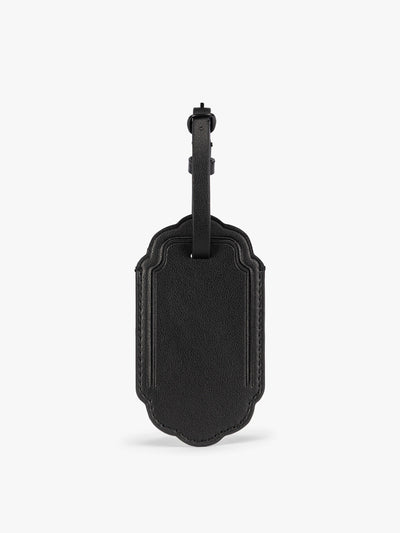 A black CALPAK Vault luggage tag with a scalloped border and a matching black strap, shown against a plain white background; ATV2601-BLACK