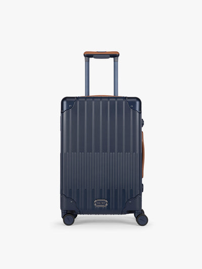 CALPAK Vault Navy blue hard-shell spinner Carry On Luggage with a telescoping handle extended, featuring a brown leatherette side handle and trim, set against a plain light gray background; LVT1020-NAVY