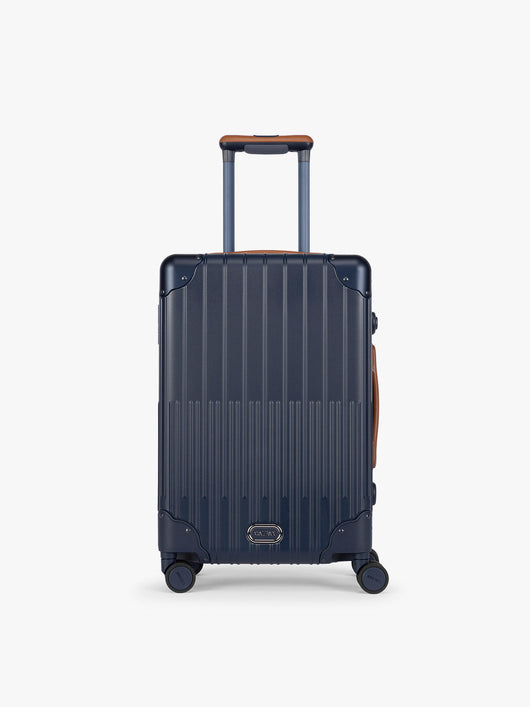CALPAK Vault Navy blue hard-shell spinner Carry On Luggage with a telescoping handle extended, featuring a brown leatherette side handle and trim, set against a plain light gray background; LVT1020-NAVY